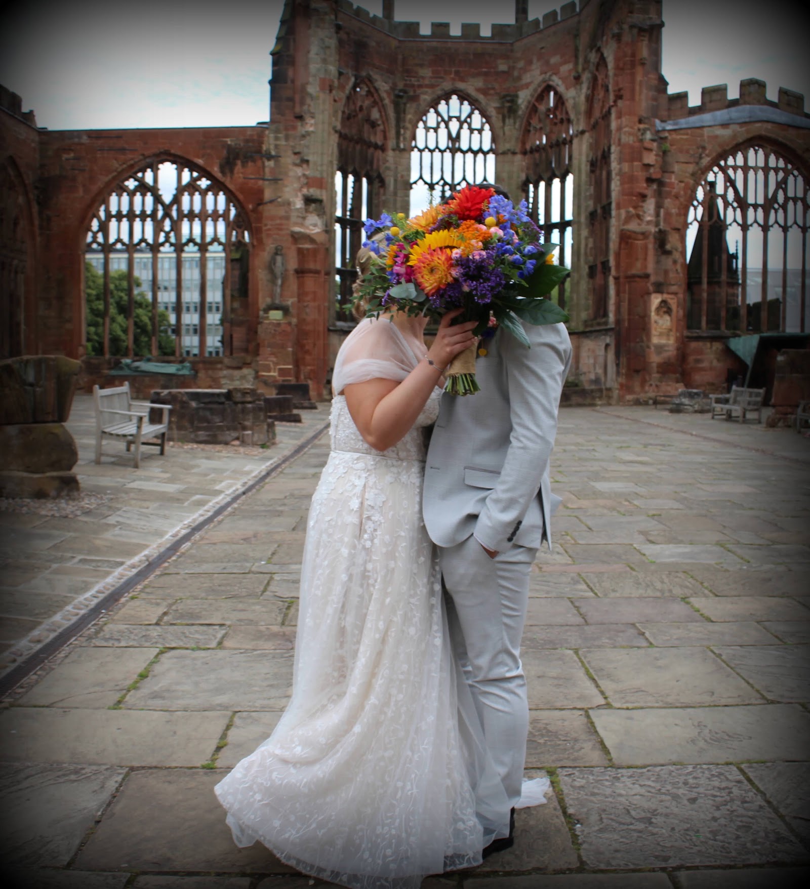 A bride and groom kissing behind a bright wildflower bouquet in the Coventry Cathedral ruins